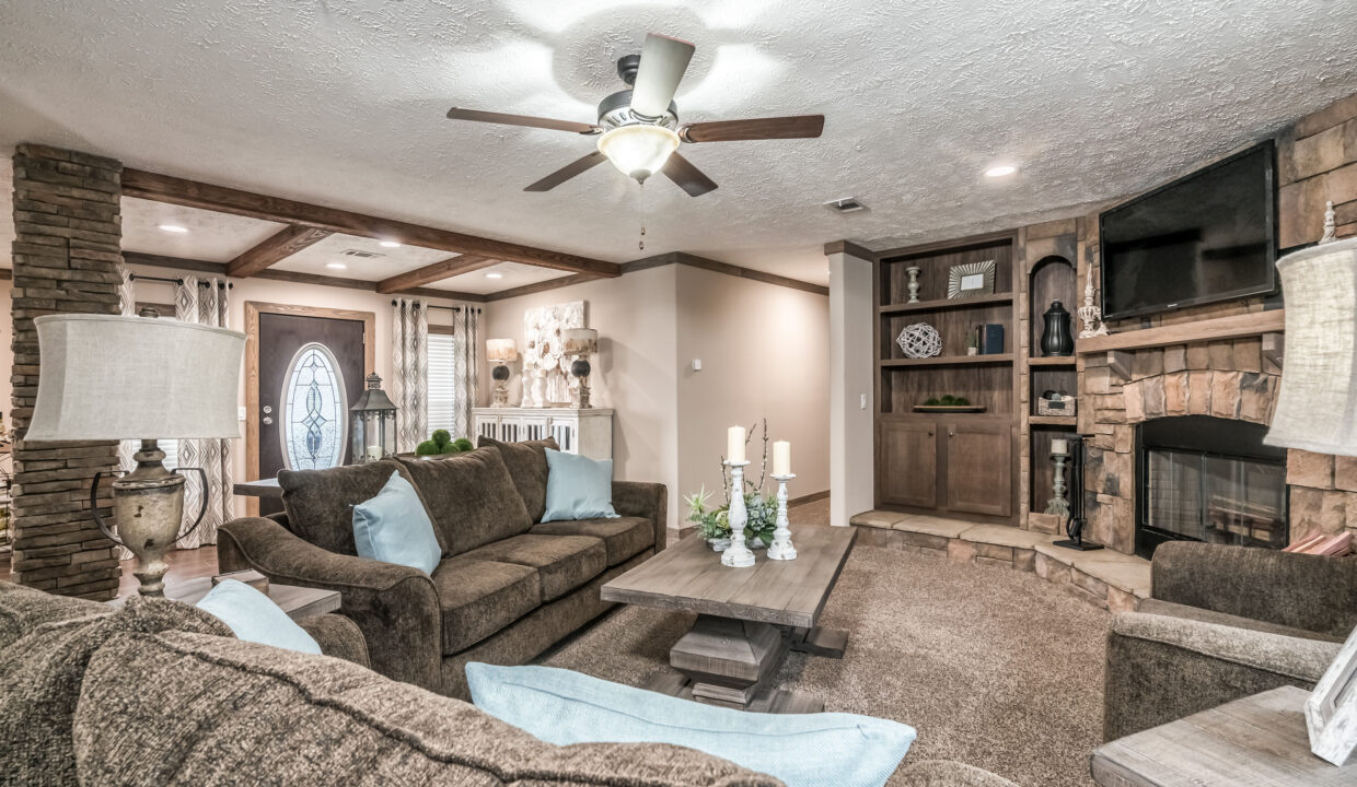 A cozy living room features a brown sectional sofa, light blue cushions, stone fireplace with TV, wooden beams, ceiling fan, and soft lighting.