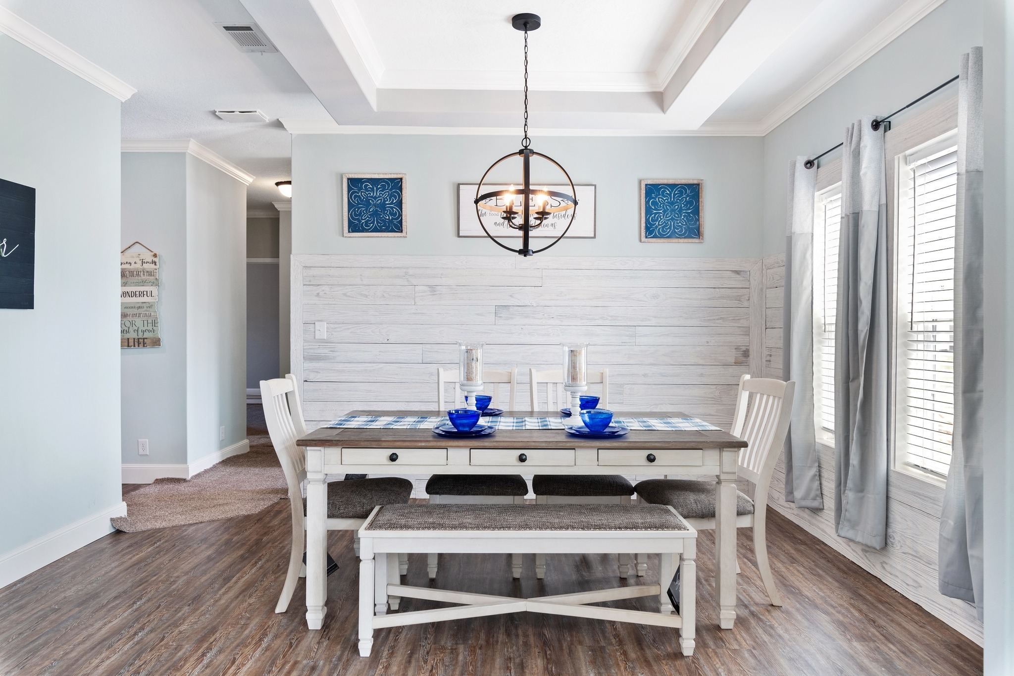 Bright dining room with a white wooden table set, blue accents, and a modern chandelier. Large windows with gray curtains add natural light.