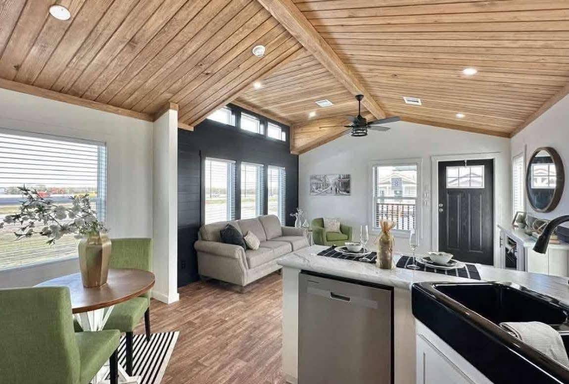 Modern living room with vaulted wooden ceiling and bright lighting. Features gray sofa, black door, green chairs, and small dining table. Cozy ambiance.