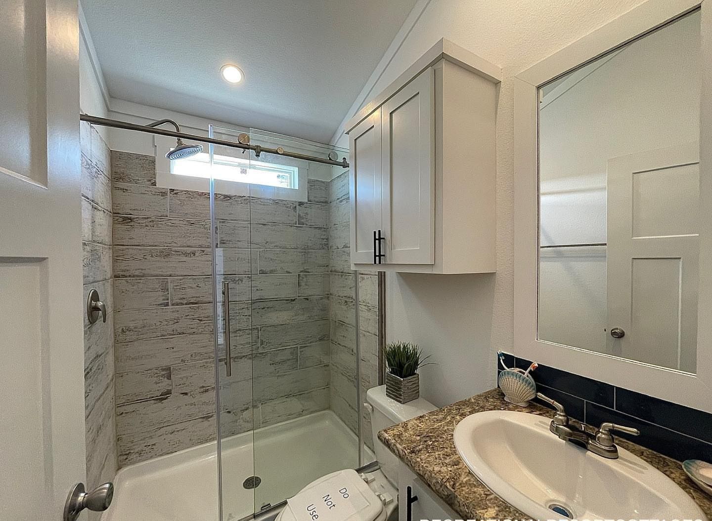 Modern, compact bathroom with a glass-enclosed shower featuring wood-look tiles. There’s a white cabinet above the toilet and a granite countertop with a sink.