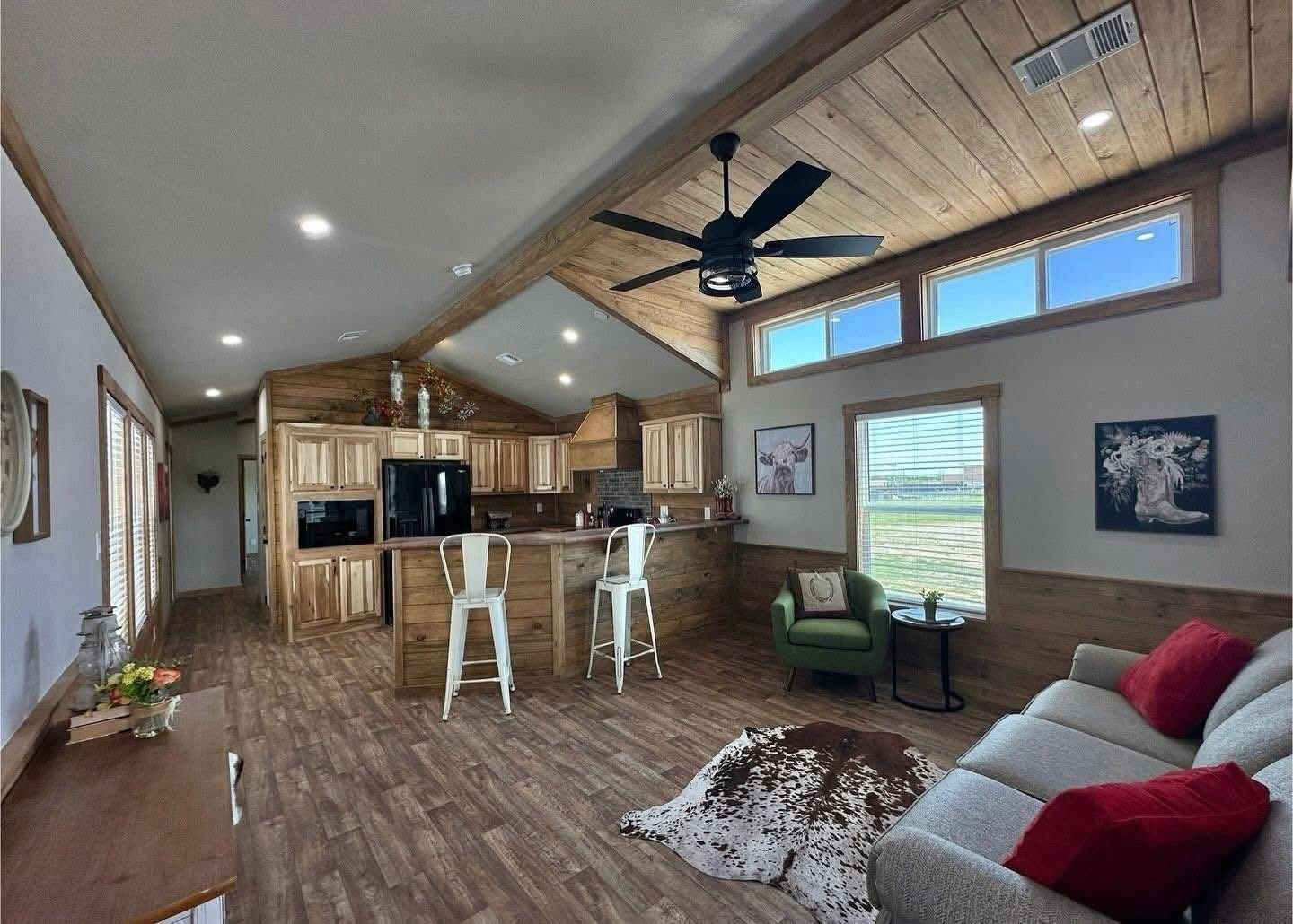 Rustic open-plan living area with wooden accents, featuring a cozy sofa with red cushions, bar stools, a cowhide rug, and large windows.