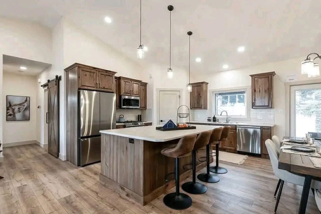 Spacious modern kitchen with wooden cabinets, large island, and four stools. Stainless steel appliances and a dining table set nearby. Bright and airy.