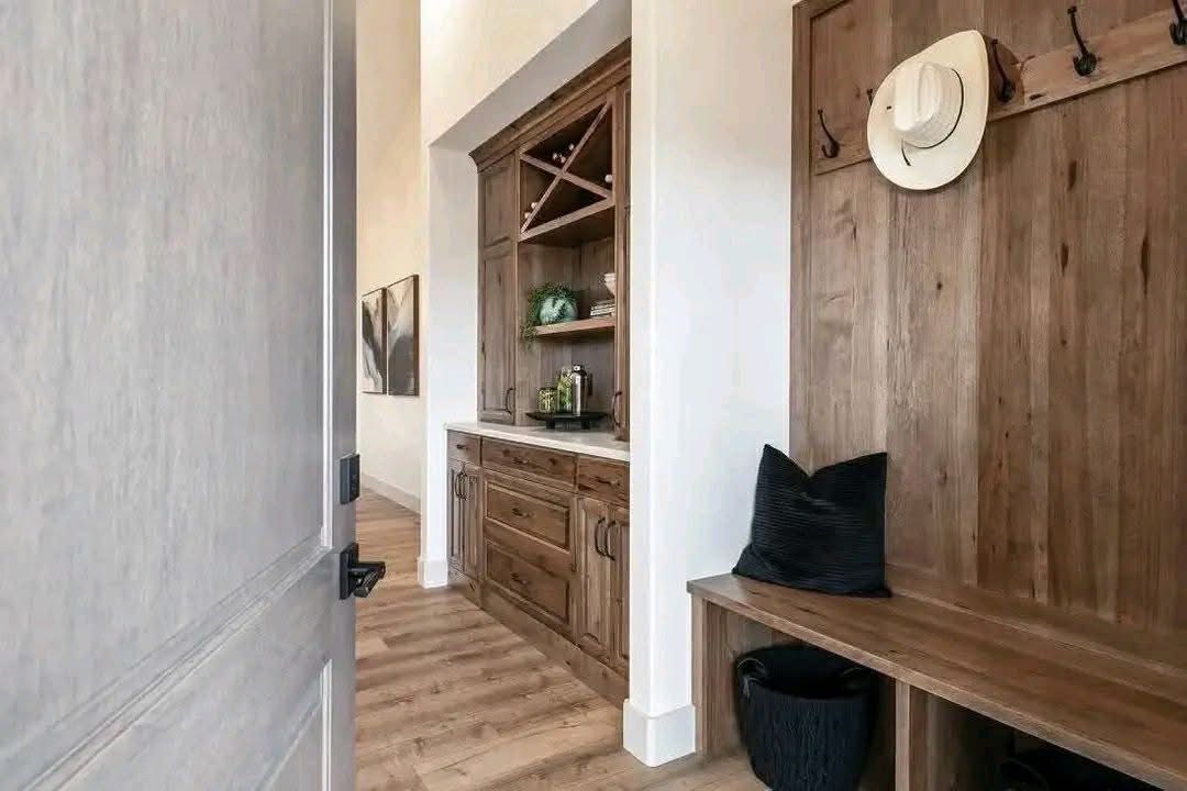 A wooden hallway features a built-in cabinet with drawers and shelves, displaying decor. A bench with a black pillow and hat hooks adds warmth.