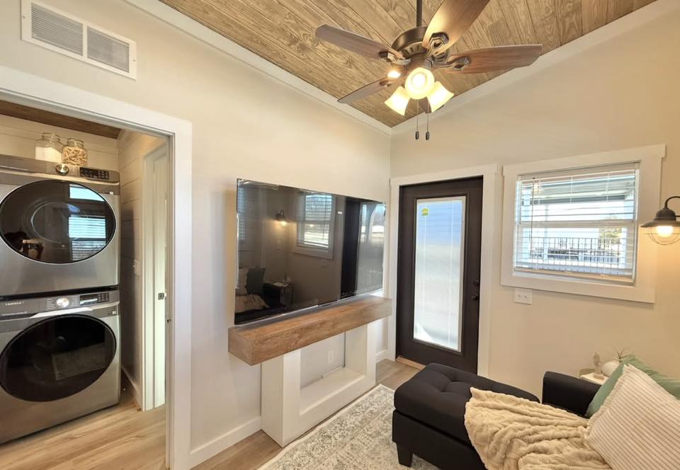 Compact laundry room with stacked washer-dryer; adjacent cozy living area with large TV, plush sofa, and rustic ceiling fan. Bright and inviting.