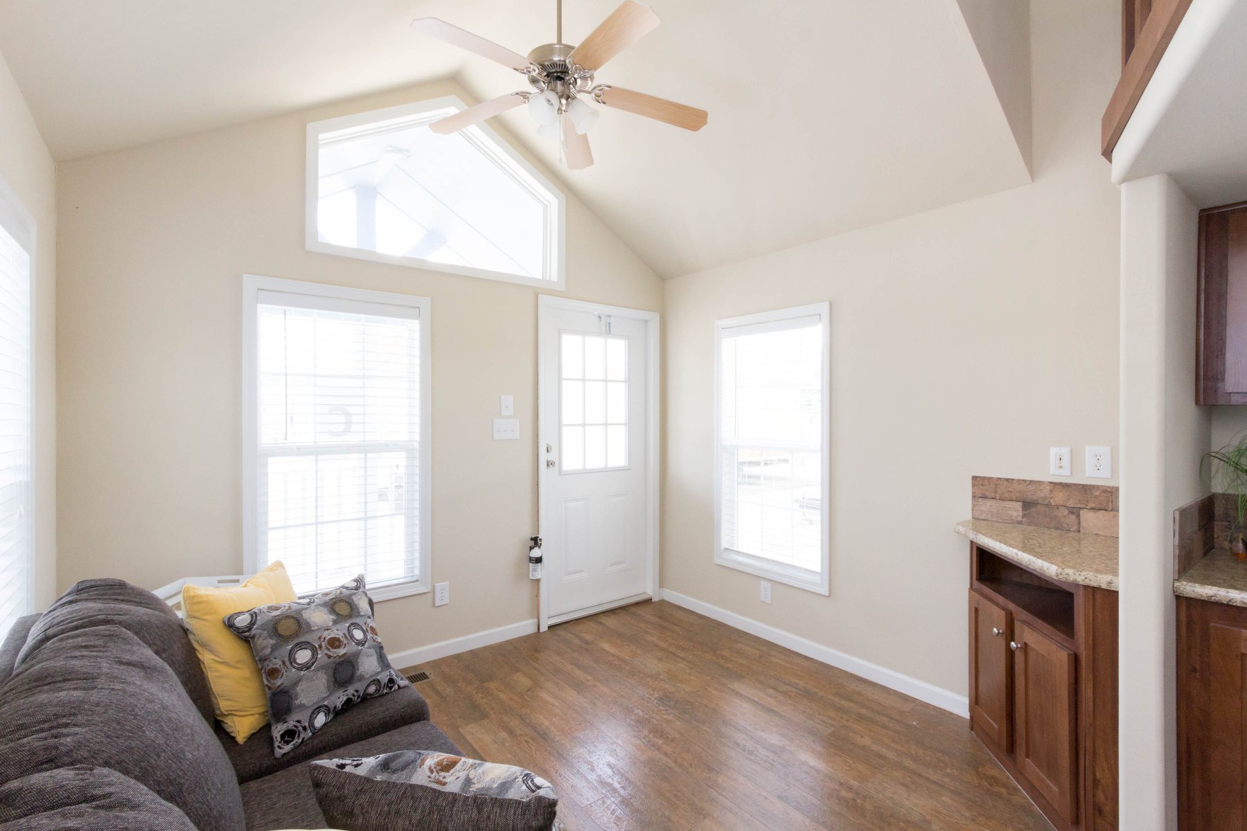 A small, bright living area with a sloped ceiling, beige walls, and wooden floors. It features a gray sofa with colorful cushions and a ceiling fan.