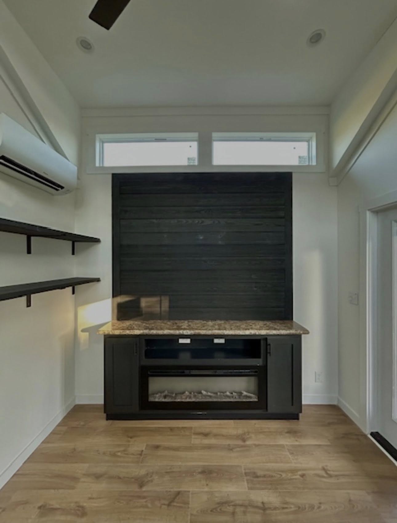 Modern living space with a sleek black fireplace, wooden mantle, floating shelves, and minimalist decor. Sunlight filters in, enhancing the cozy atmosphere.