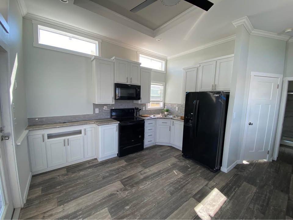 Bright kitchen with white cabinets, black appliances, and light wood flooring. Sunlight streams through upper windows, creating a warm, inviting atmosphere.