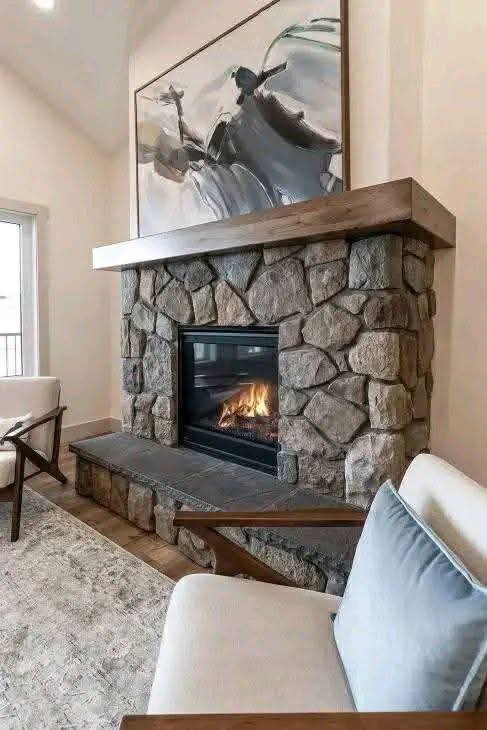 Cozy living room with a stone fireplace, warm flames, and modern art above. Soft chairs and natural tones create a welcoming and serene atmosphere.