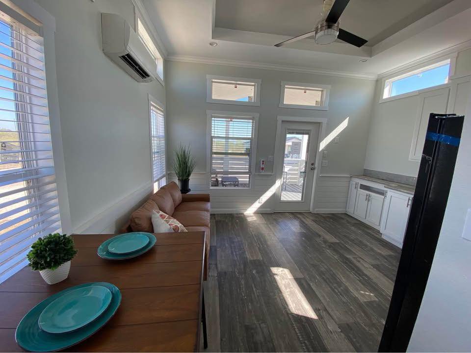 Bright, modern living space with wooden floors, a brown sofa, turquoise plates on a table, and white cabinets. Sunlight streams through large windows.
