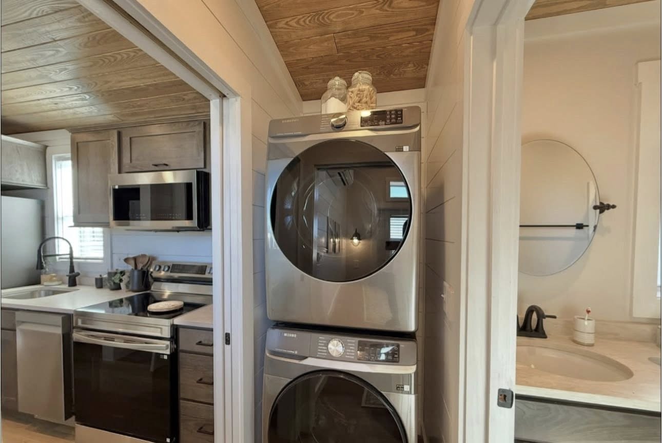 A compact laundry nook with a modern, gray stacked washer and dryer is flanked by a cozy kitchen and a bathroom. The kitchen features wooden cabinets, a stainless steel fridge, and a microwave, while the bathroom shows a round mirror above a countertop. The atmosphere is warm and organized.