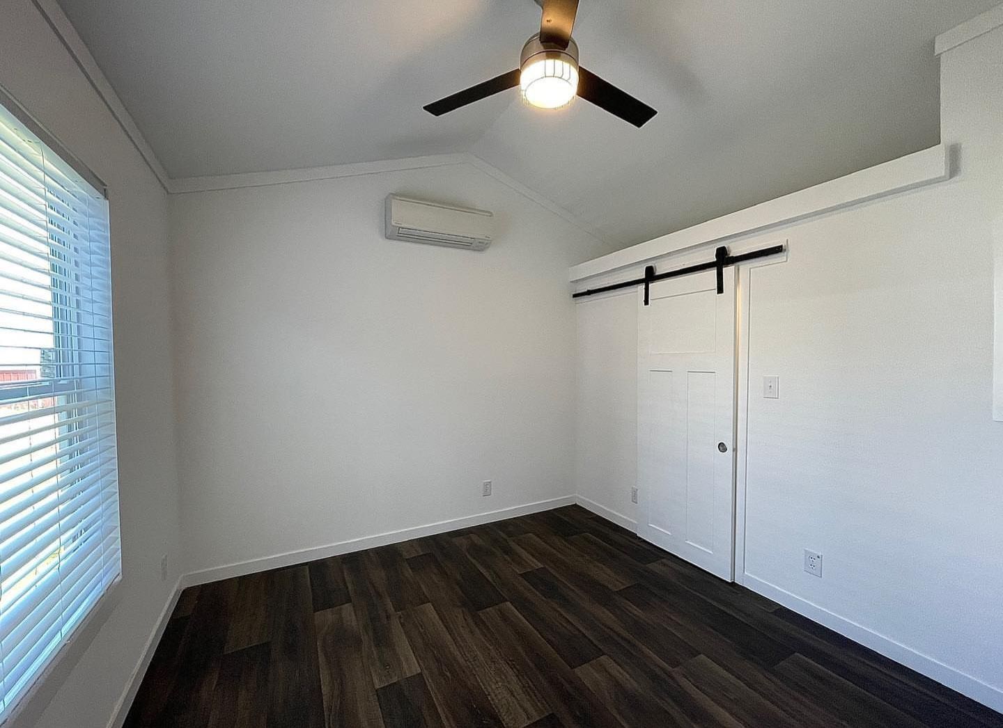 A minimalist room with dark wood floors, white walls, a sliding barn door, and a ceiling fan. Natural light filters through blinds, creating a calm ambiance.