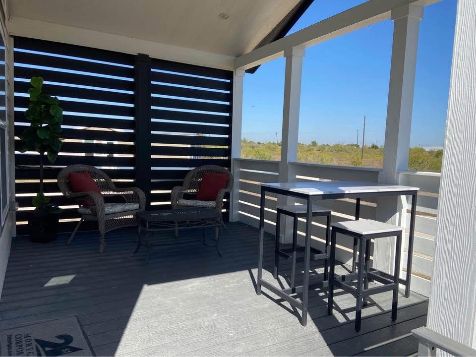 Covered patio with wicker chairs and red cushions, small table, and high-top bar table set. Sunlight filters through wooden slats, creating a serene atmosphere.
