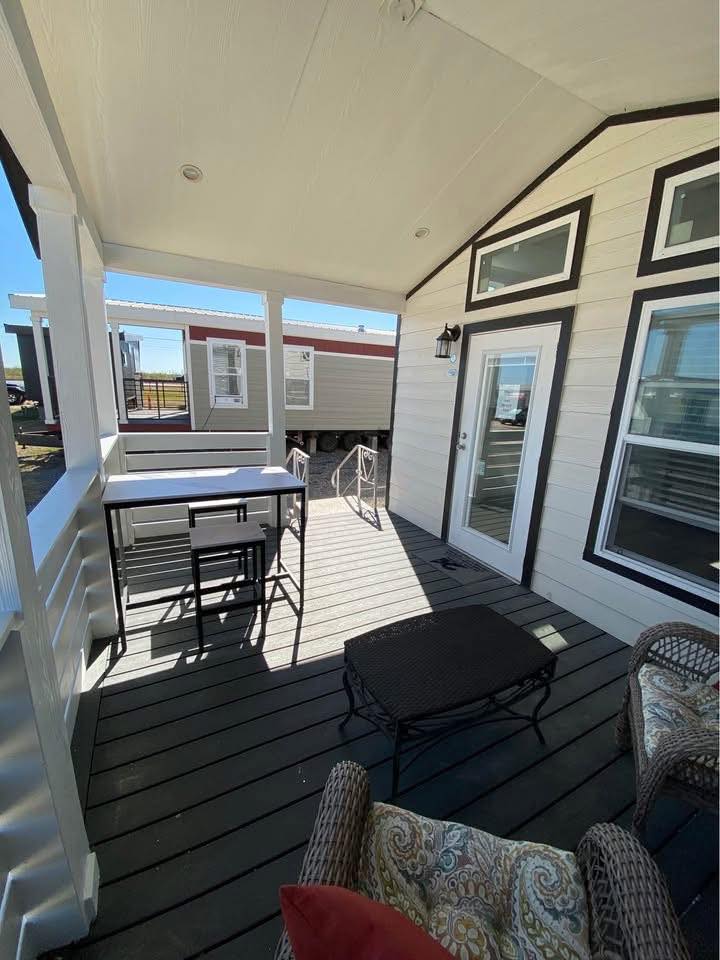 Covered porch with light gray siding and black trim. Features outdoor furniture including wicker chairs and a table. Bright, sunny day.