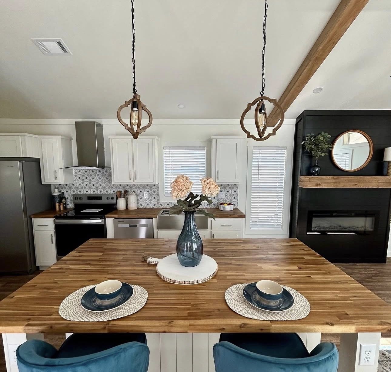 Modern kitchen with wooden island, blue velvet stools, and elegant decor. White cabinets, patterned backsplash, and pendant lights create a warm ambiance.