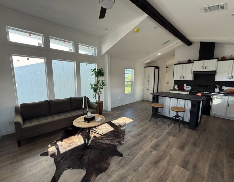 Modern living area with a gray sofa, round wooden coffee table, and potted plant. Rustic kitchen features an island with stools, white cabinets, and skylight windows. Warm, inviting atmosphere.