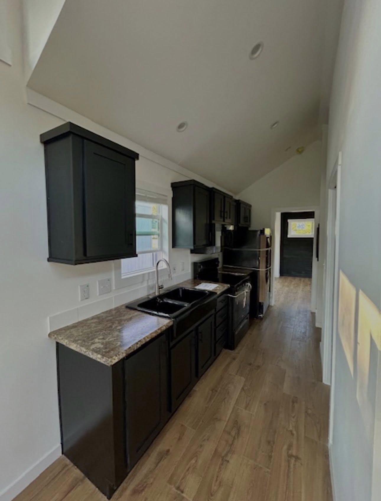Narrow kitchen with dark cabinets, wooden floor, and marble countertops. Bright window above sink; appliances line one wall. Cozy and modern feel.