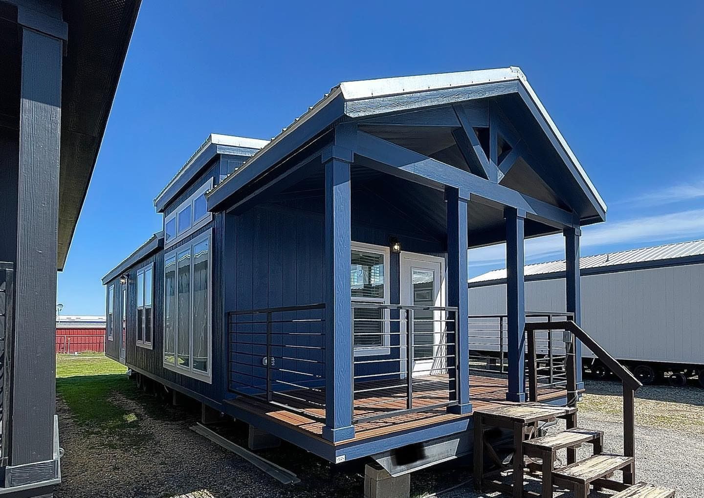 Blue tiny home with a gabled roof and wooden porch railing under a clear blue sky. Stairs lead up to a glass door, evoking a cozy, modern vibe.