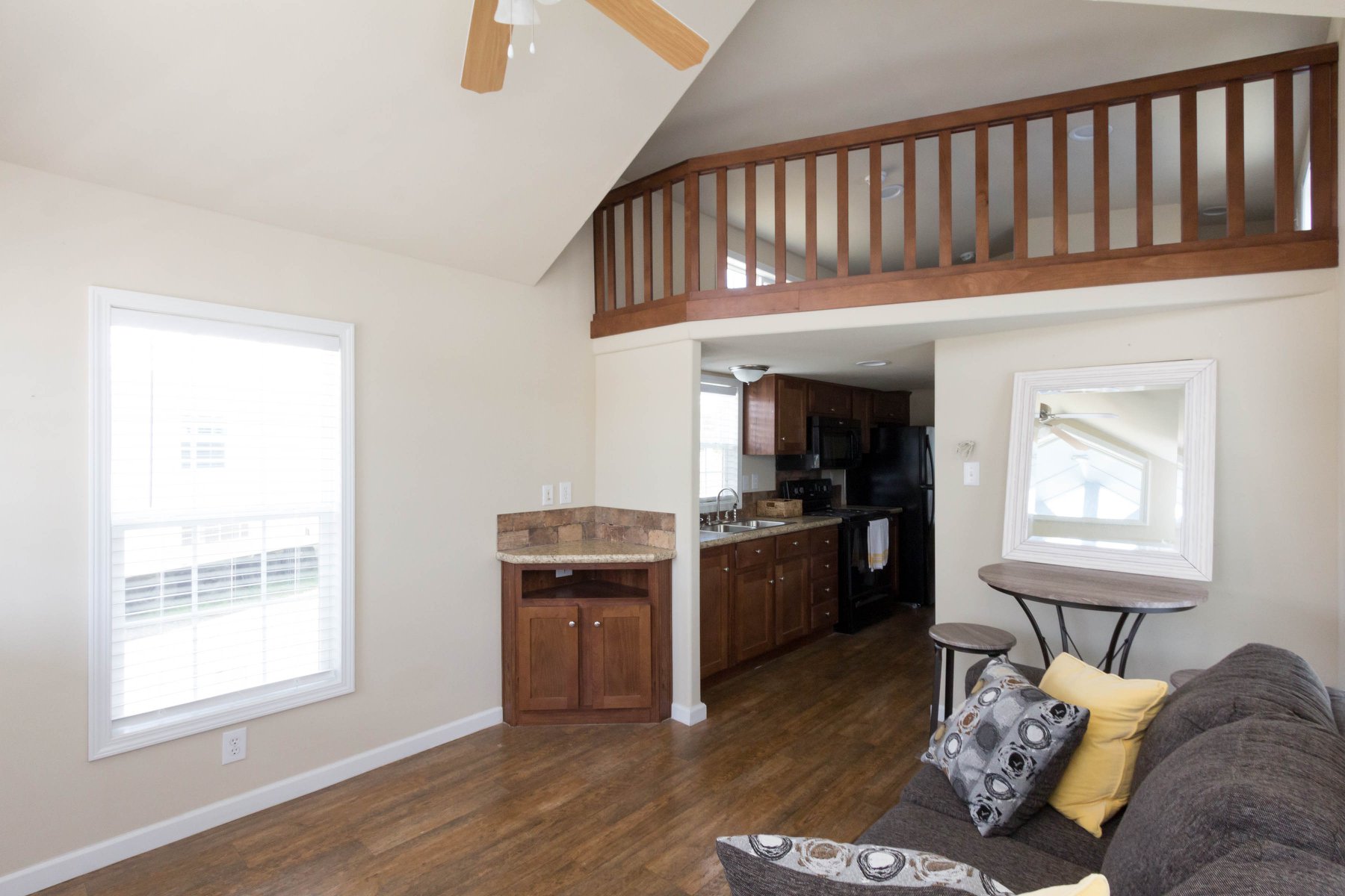 Spacious living room with vaulted ceiling and loft. Features wooden floors, a brown couch with yellow and gray pillows, open kitchen, and large window.