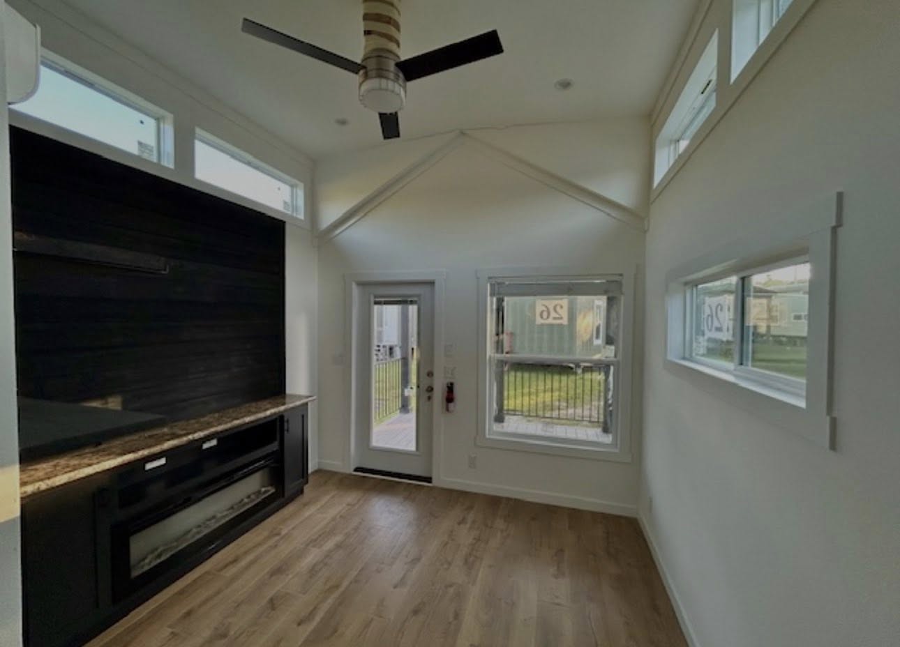 Modern tiny home interior with vaulted ceiling and light wood flooring. Features a large black fireplace, abundant natural light, and ceiling fan.