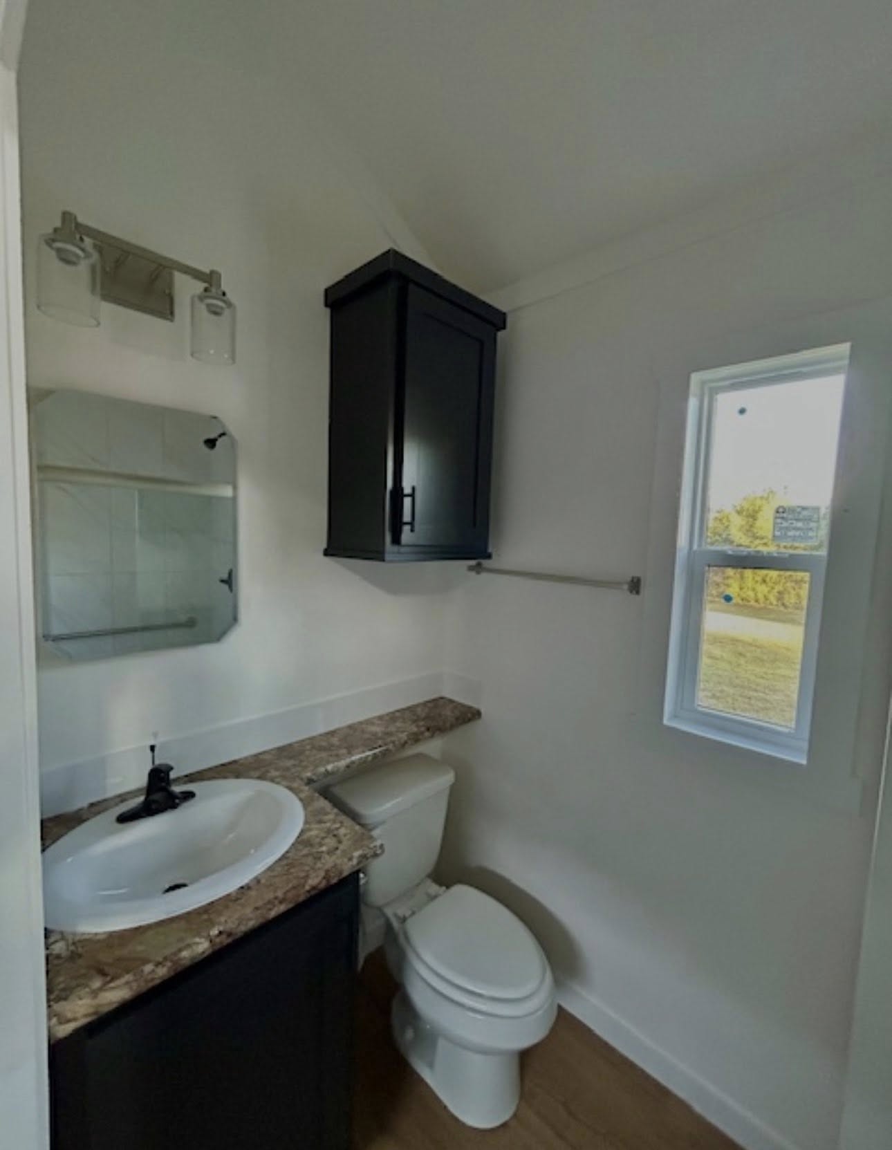 Compact bathroom with a white sink on a brown countertop, black cabinet, toilet, small window, and modern light fixture on white walls.