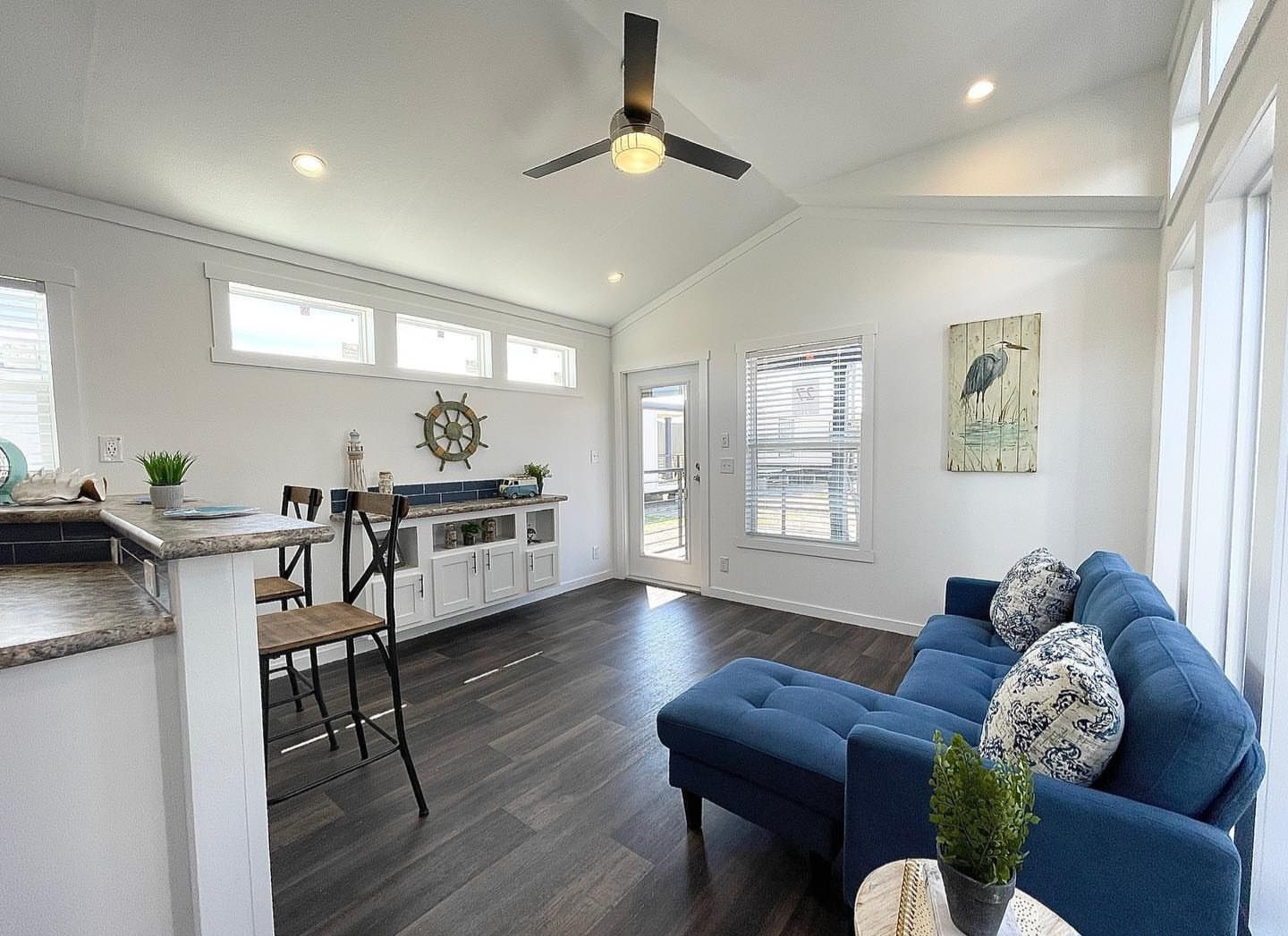 Modern living room with high ceilings, large windows, and dark wood flooring. Features a blue sofa, decorative cushions, nautical-themed decor, and a ceiling fan. Bright and inviting atmosphere.