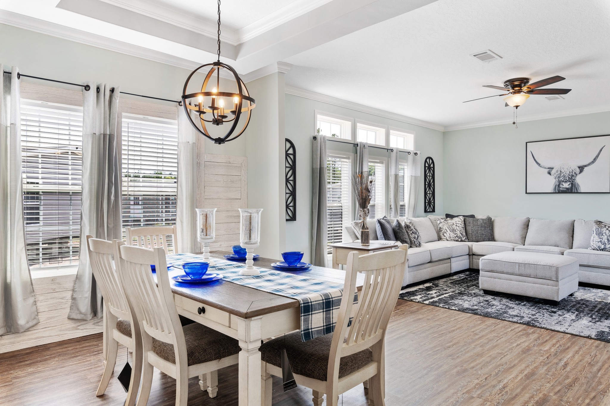 Bright, modern dining and living area. Wooden table with blue accents, beige sofa, bull artwork, large windows, and a ceiling fan evoke a cozy feel.