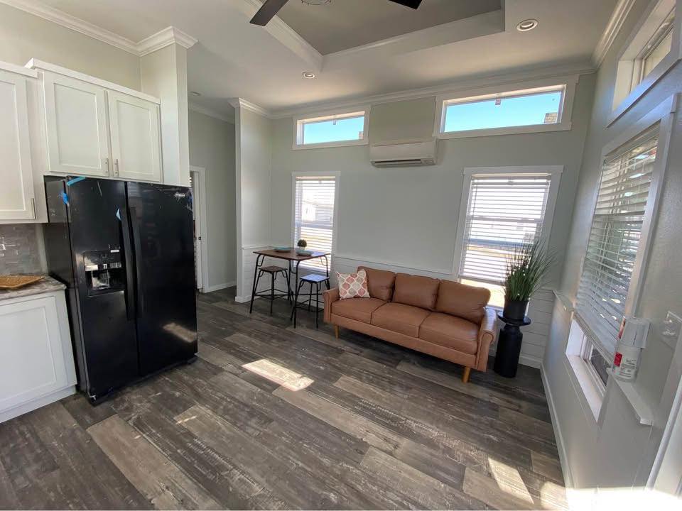 A small, bright living area with light gray walls and a wood floor. Features a black fridge, tan sofa, bar table, and large windows with ample daylight.