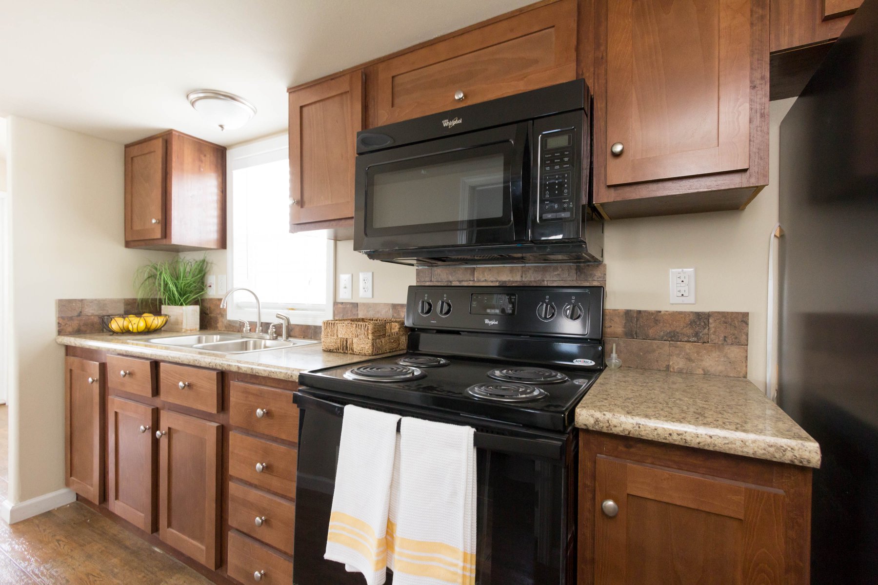 Warm kitchen with wood cabinets, black appliances, and granite countertops. A white towel hangs on the stove. Natural light from a window enhances the cozy feel.