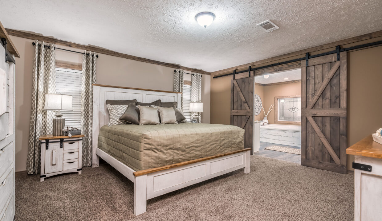 A cozy bedroom features a large bed with a neutral green quilt, flanked by two nightstands with lamps. Barn doors open to a glimpse of a bathroom, enhancing the rustic charm.