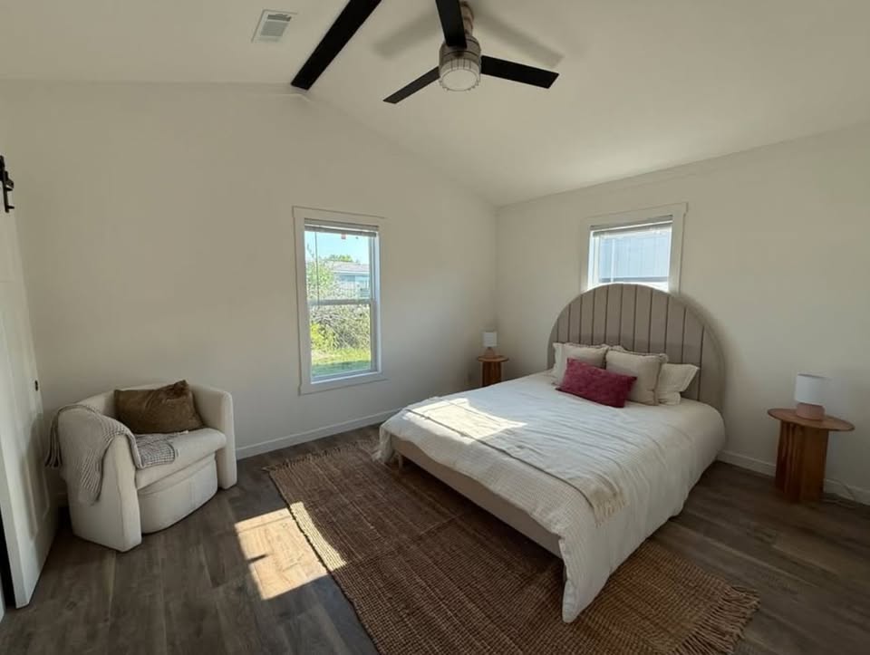 Minimalist bedroom with a neatly made bed, wooden headboard, and side tables. A cozy chair sits by the window. Soft lighting creates a calm ambiance.