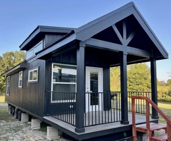 Black modern tiny house with large windows and a porch set on concrete blocks. Surrounded by green trees, clear blue sky, creating a peaceful vibe.