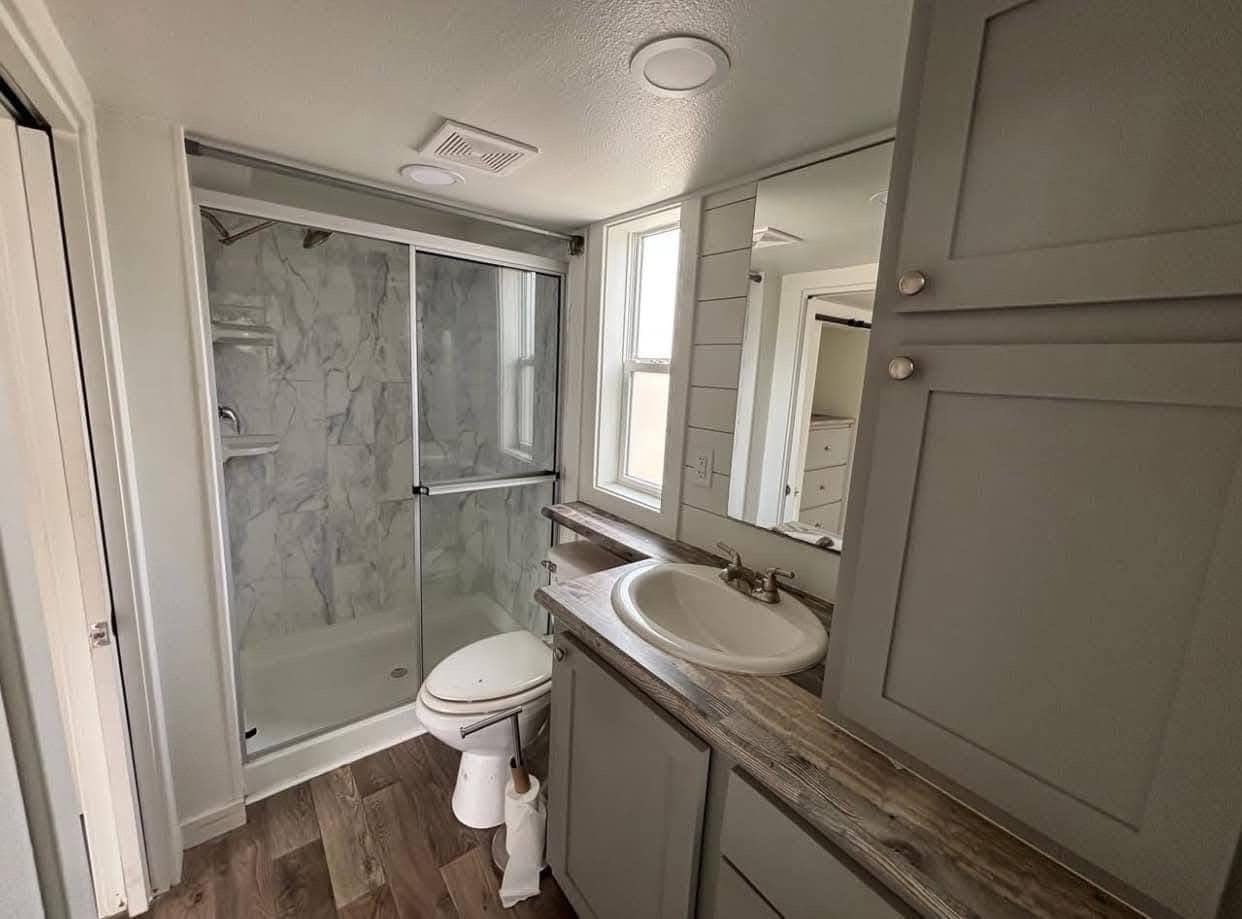 Compact, modern bathroom with gray cabinetry, a wooden countertop, sink, and mirror. Marble-tiled shower with glass doors and wood-patterned floor.