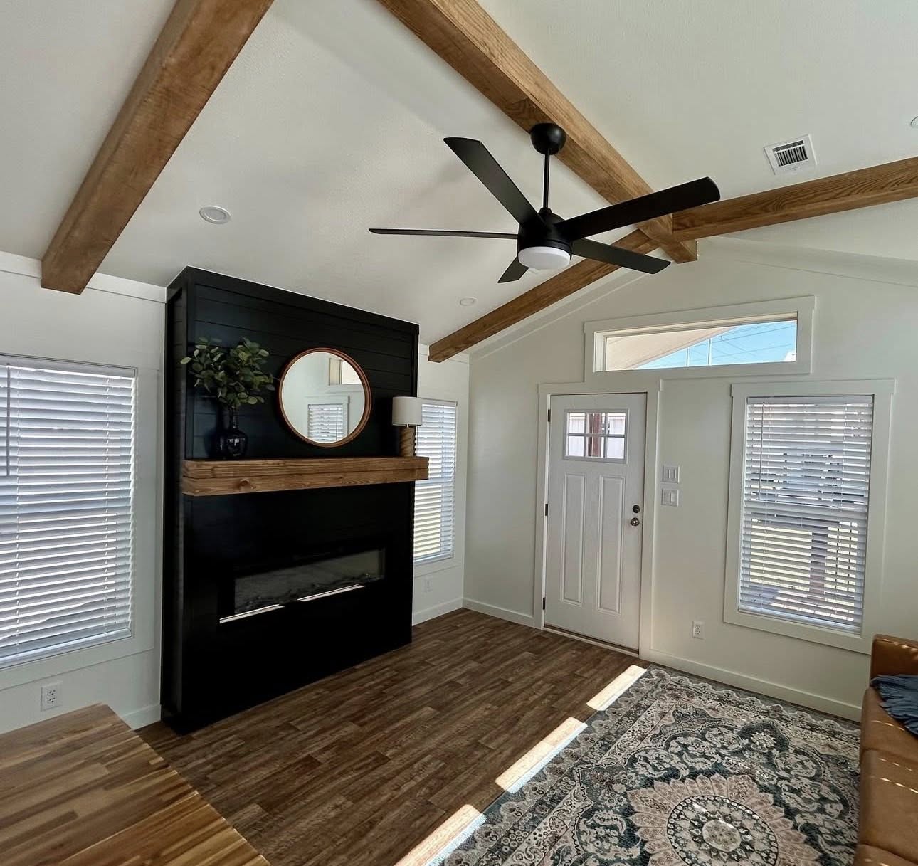 Cozy living room with wooden beams and fan above. Features a modern black fireplace, round mirror, and plants. Sunlight streams through large windows.