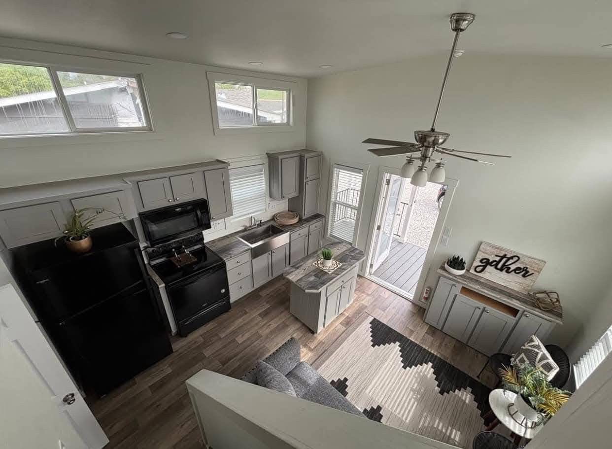 Modern open-plan kitchen and living area viewed from above. Gray cabinets, black appliances, a small island, cozy seating, and natural light create a welcoming space.