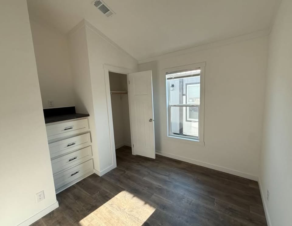 Spacious empty room with white walls, wood flooring, and sunlight through a window. Features an open closet and a dresser with black countertop. Minimalist vibe.