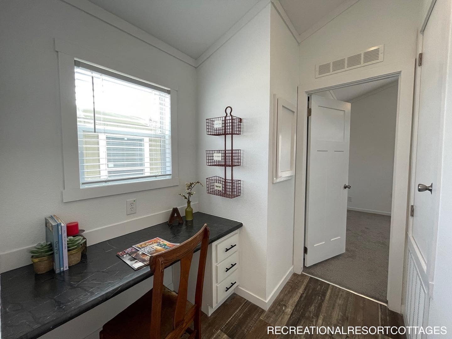 Compact home office nook with a dark countertop, wooden chair, books, magazines, and decor. A window provides natural light, emitting an inviting, cozy vibe.