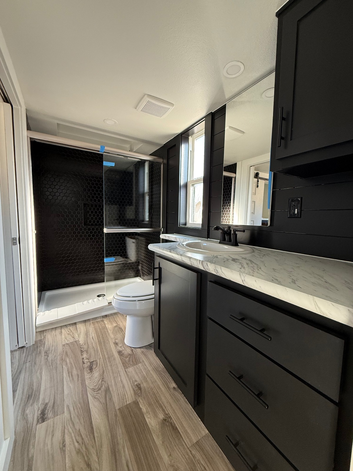Modern bathroom featuring dark cabinetry and black hexagonal tiles. Marble countertop with dual sinks, wood-look flooring, and a glass shower door.