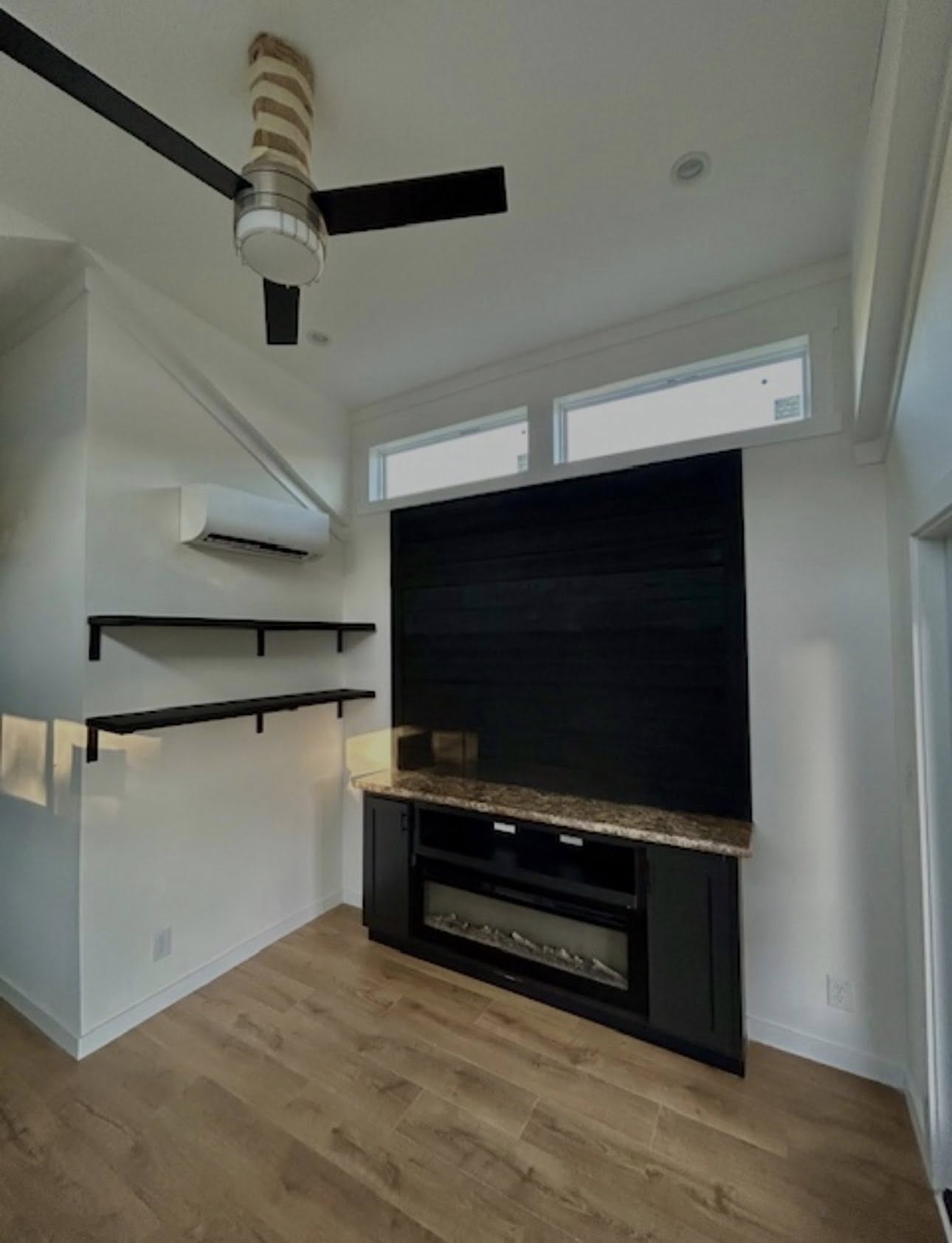 Modern room corner with wood flooring, ceiling fan, and black shelving under an air conditioner. A fireplace with a dark surround adds warmth.