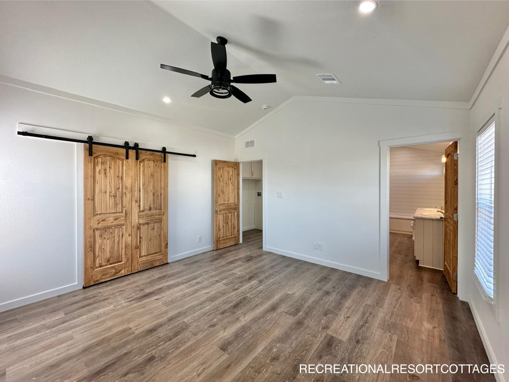 A spacious, empty room with wood flooring and white walls. It features rustic wooden barn doors, a ceiling fan, and abundant natural light, creating a warm, inviting atmosphere.