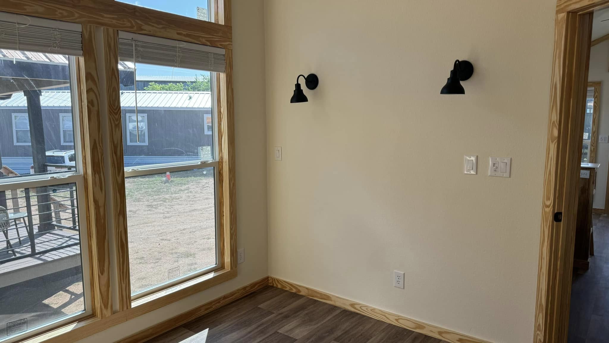 Bright room with large, sunlit windows featuring wood trim. The walls are light-colored, with two black wall sconces. Wooden floor adds warmth.