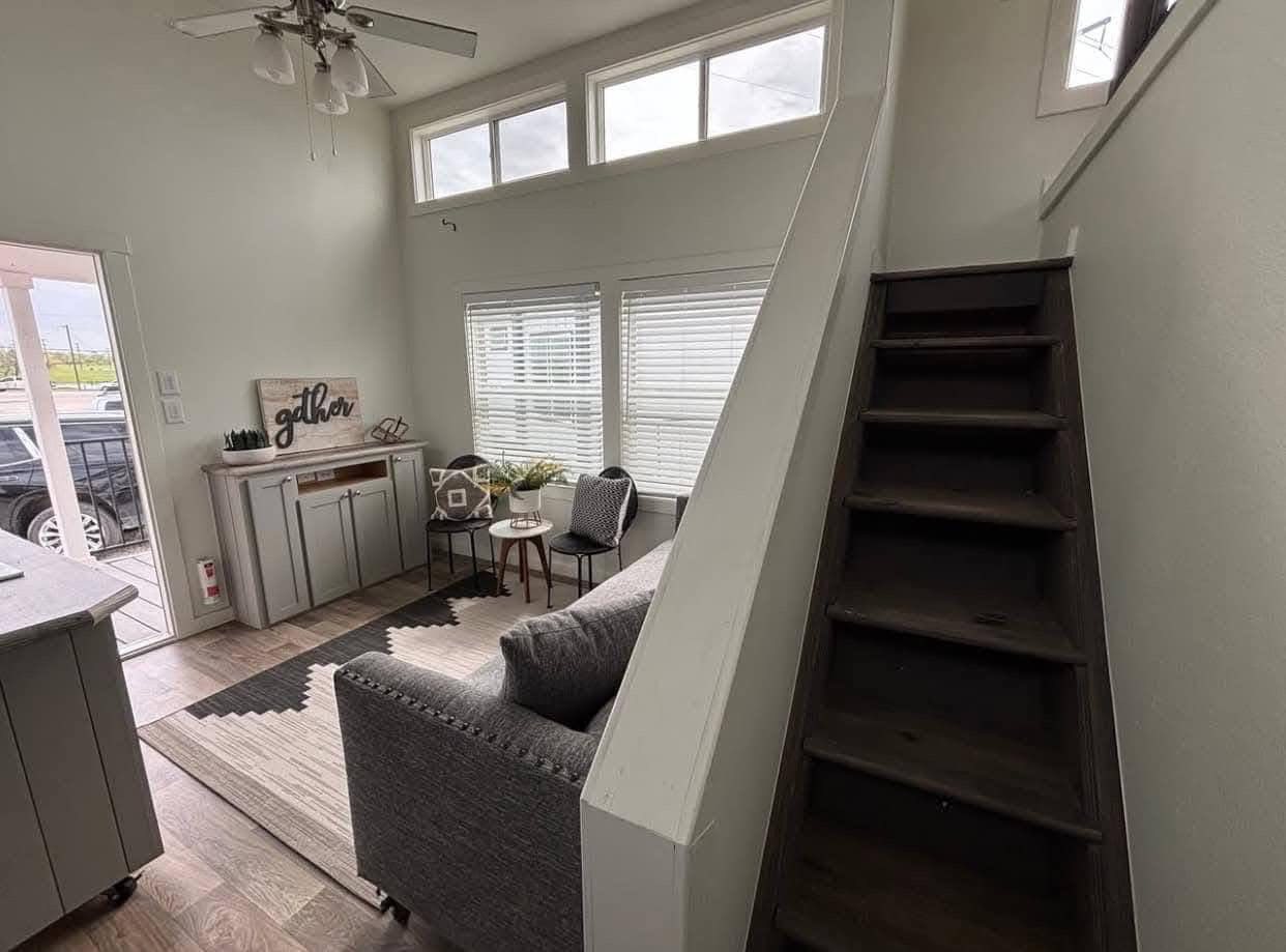 Cozy living room with a gray sofa, small dining table with chairs, and stairs leading up. Large windows and a sign reading "gather" add warmth. Ceiling fan above.