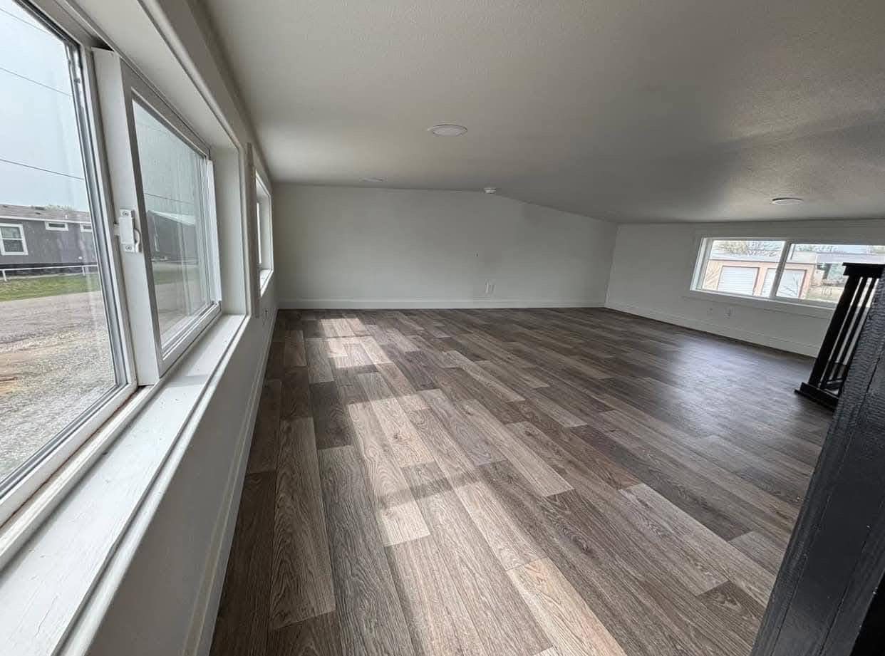 Spacious room with light gray walls and wide plank wood flooring. Large windows with natural light create a bright, airy atmosphere. Minimalist design.