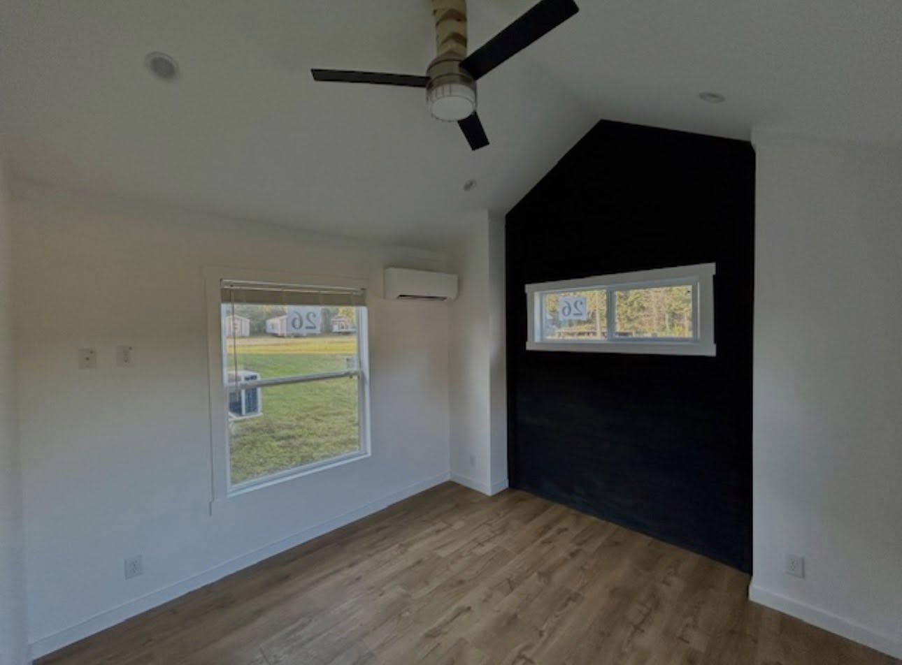 Minimalist room with wood flooring, a black accent wall, and two windows letting in natural light. Ceiling fan and air conditioner present.