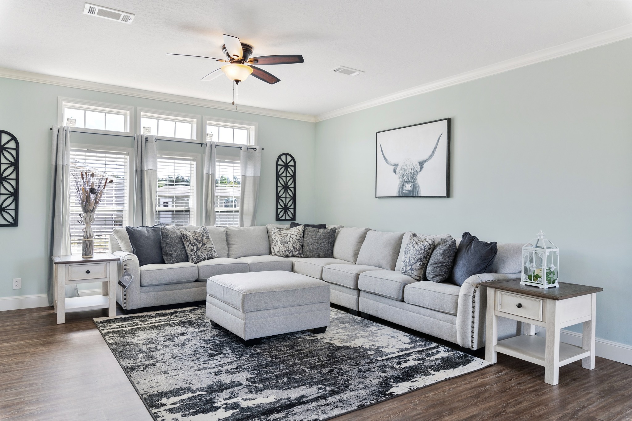 A modern living room with a large gray sectional sofa, patterned cushions, a central ottoman, and a monochrome rug. A bull art piece hangs on pale blue walls. Natural light filters through three windows with curtains. A ceiling fan adds a cozy touch.
