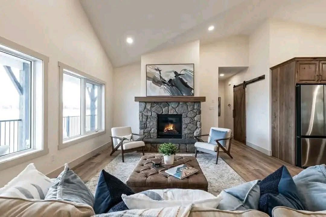 A cozy living room with a stone fireplace, modern artwork above, and two white chairs. Large windows on the left, and a tufted ottoman in front.