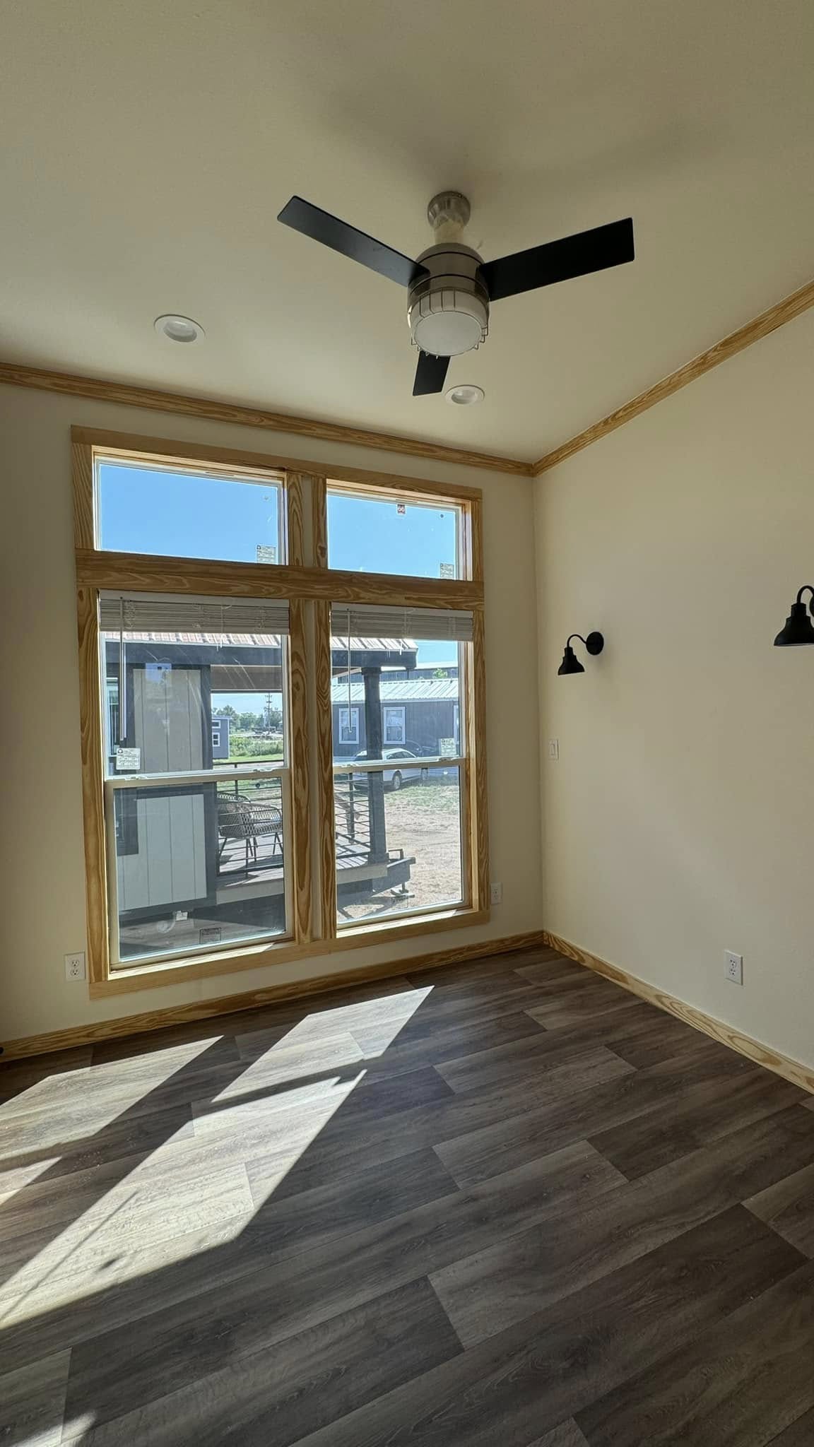A small, empty room with wood floors, large windows, and a ceiling fan. Sunlight streams through, casting shadows on the floor. Black wall sconces adorn the walls.