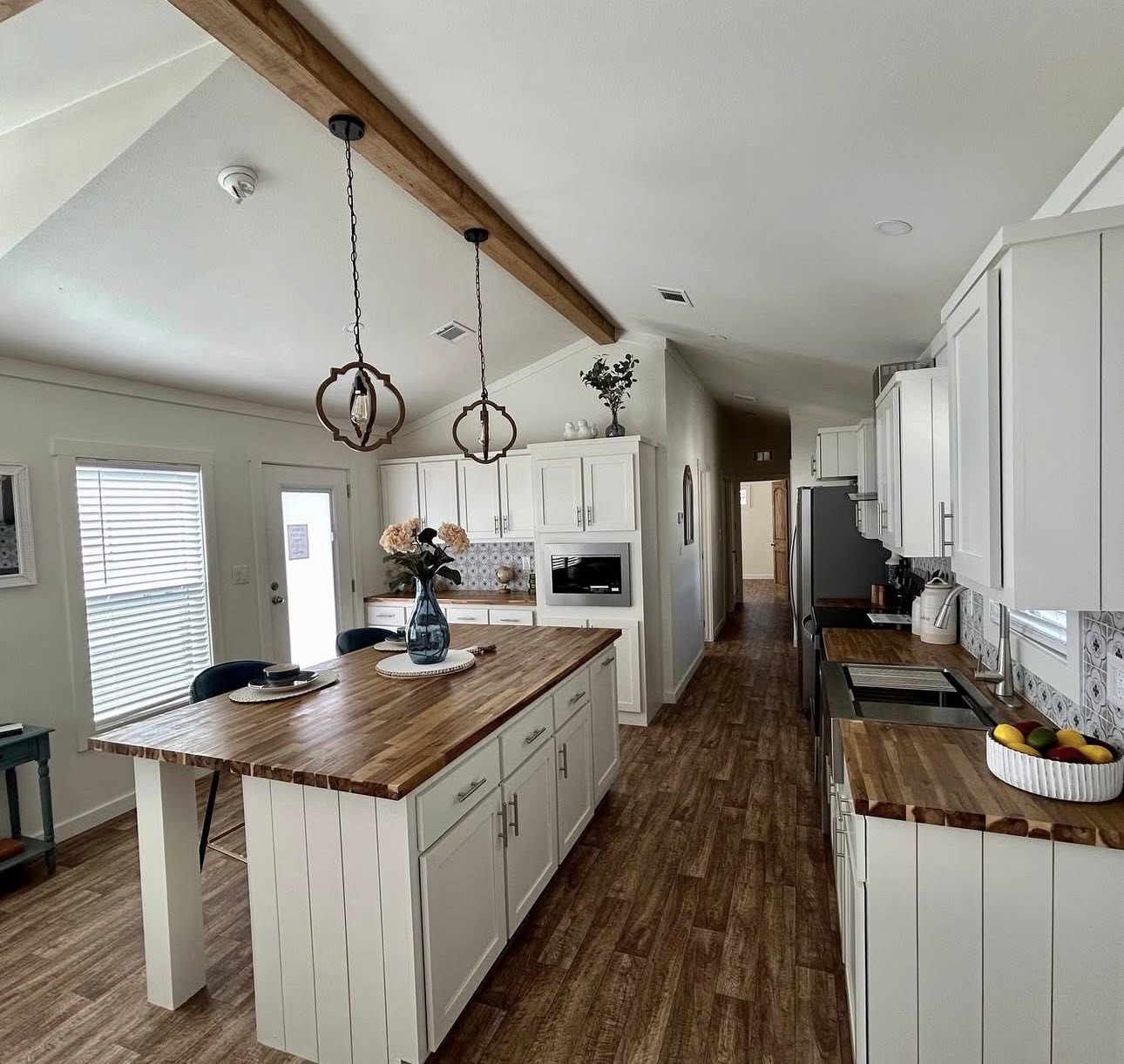 Bright kitchen with white cabinets, wood countertops, and island center. Two pendant lights hang from a wooden beam. Warm, inviting atmosphere.