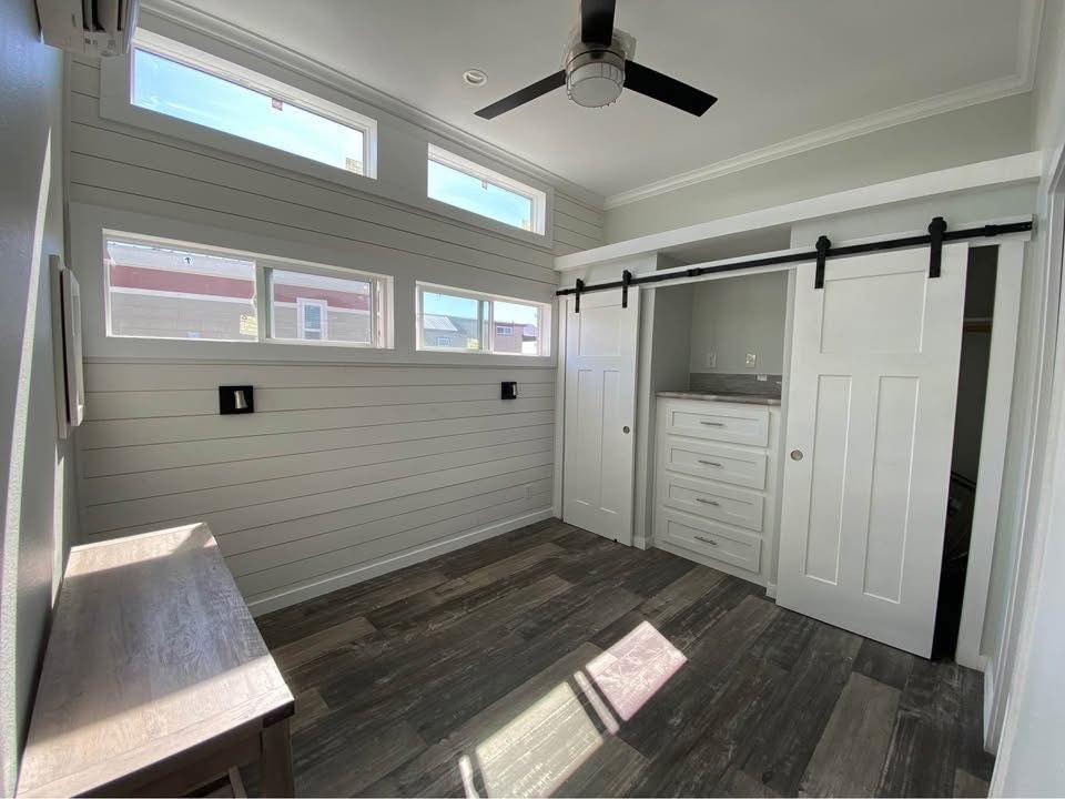 Bright, modern room with white shiplap walls, tall windows, and dark wood floor. Features sliding barn doors on closet and a ceiling fan above.