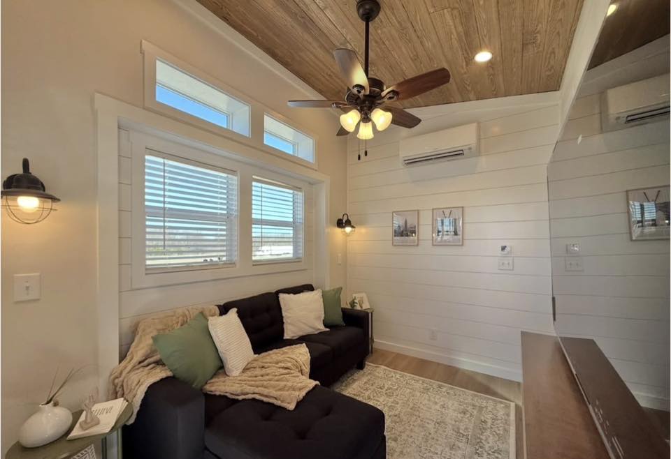 Cozy living room with a navy sofa, white and green cushions, and a beige throw. Wood ceiling with fan, shiplap walls, and large windows create an airy feel.