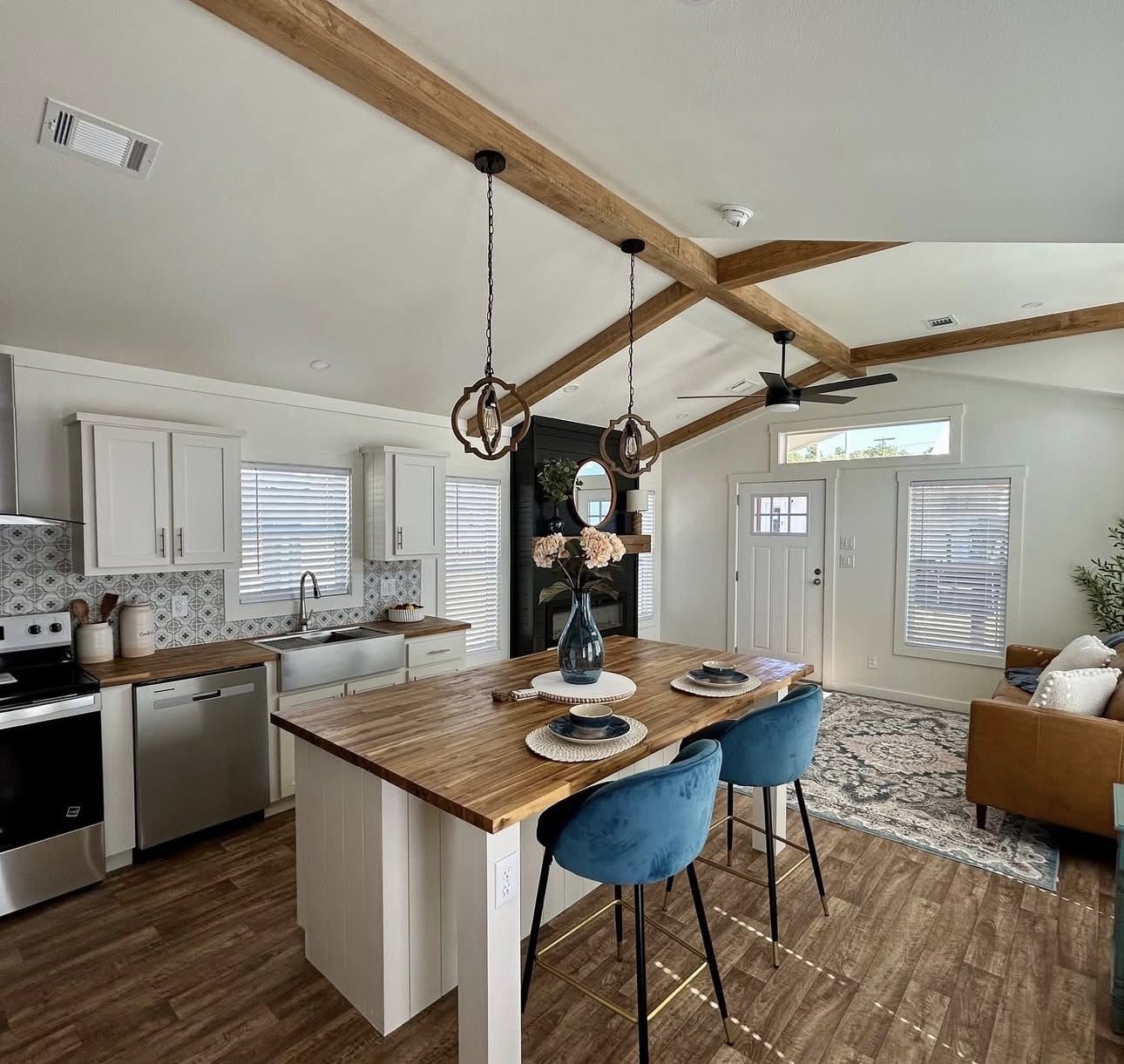Stylish open-concept kitchen and living area with wood beams, blue barstools at an island, and cozy sofa. Light streams through windows, creating a warm, inviting ambiance.